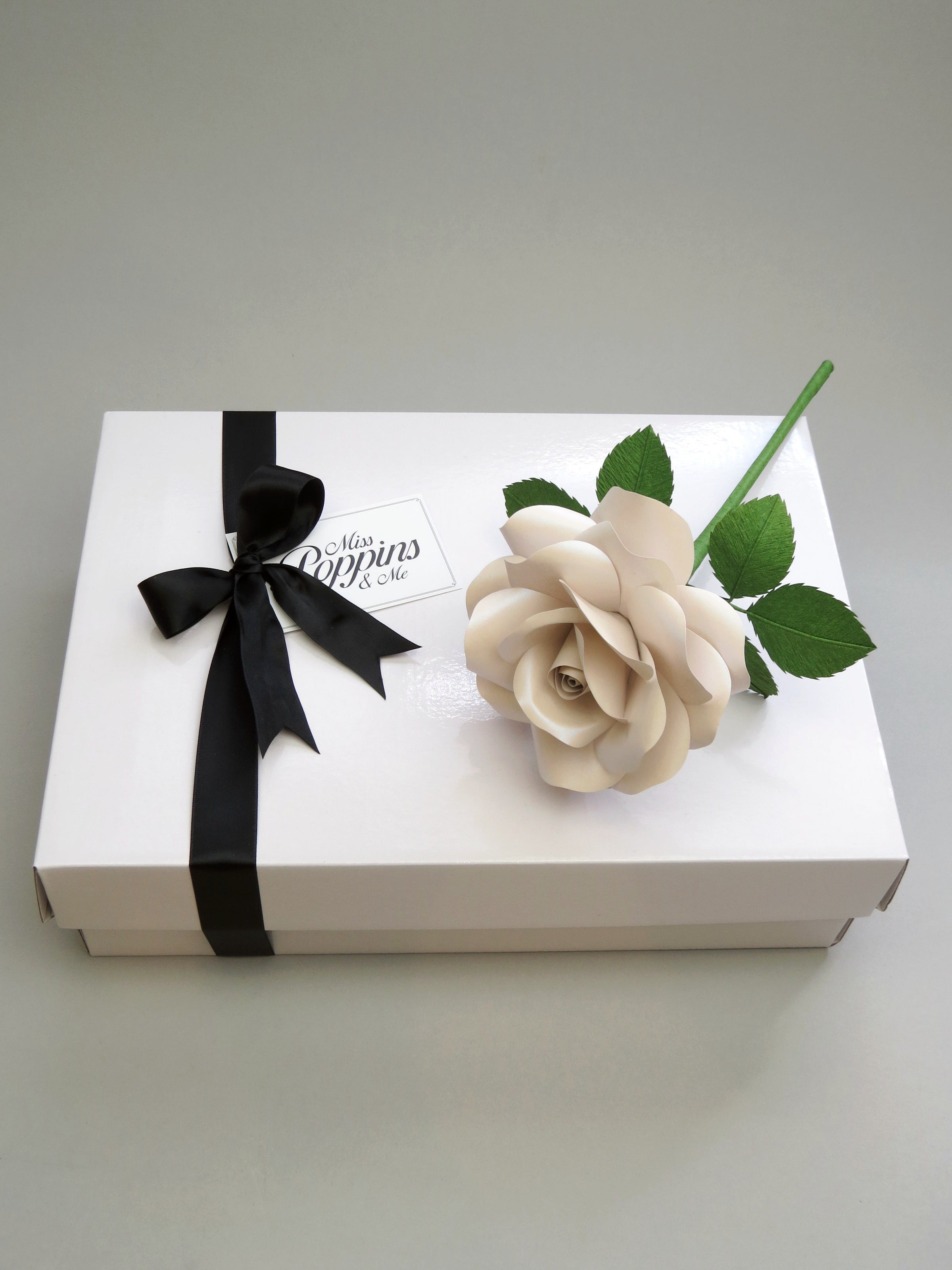 Shimmery pearl paper rose lying diagonally on top of a luxury white gift box that has a black satin ribbon tied in a bow with a Miss Poppins and Me gift tag attached