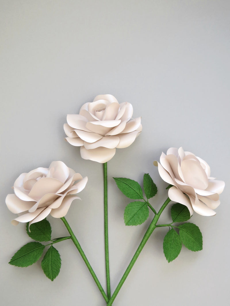 Three shimmery pearl paper roses randomly lying next to each other on a light grey background. The left rose has three ivy green leaves attached, the middle rose is leafless, the middle and the right rose has six ivy green leaves.