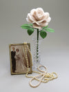 Pearl paper rose with six ivy green leaves standing in a slender glass vase to the right of a vintage gold photo frame with a sepia photo of a happy vintage bride and groom, with a long stranded pearl necklace delicately laid over the right corner of the frame and sprawled across the foreground of the photo