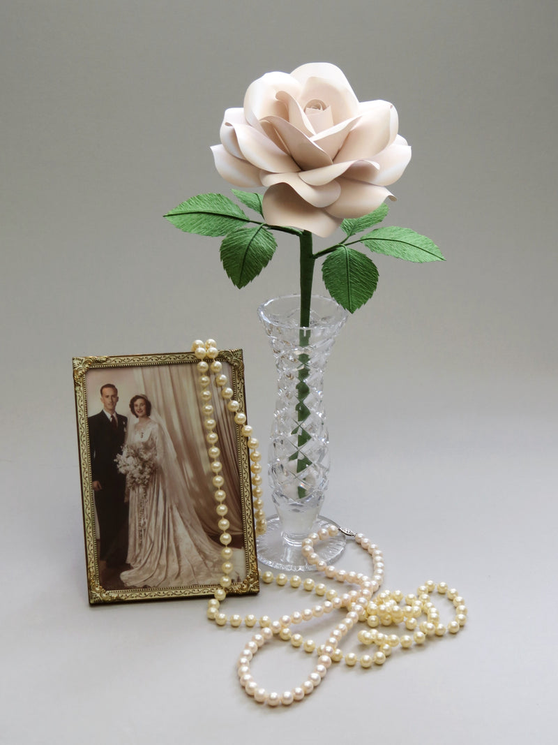 Pearl paper rose with six ivy green leaves standing in a slender glass vase to the right of a vintage gold photo frame with a sepia photo of a happy vintage bride and groom, with a long stranded pearl necklace delicately laid over the right corner of the frame and sprawled across the foreground of the photo