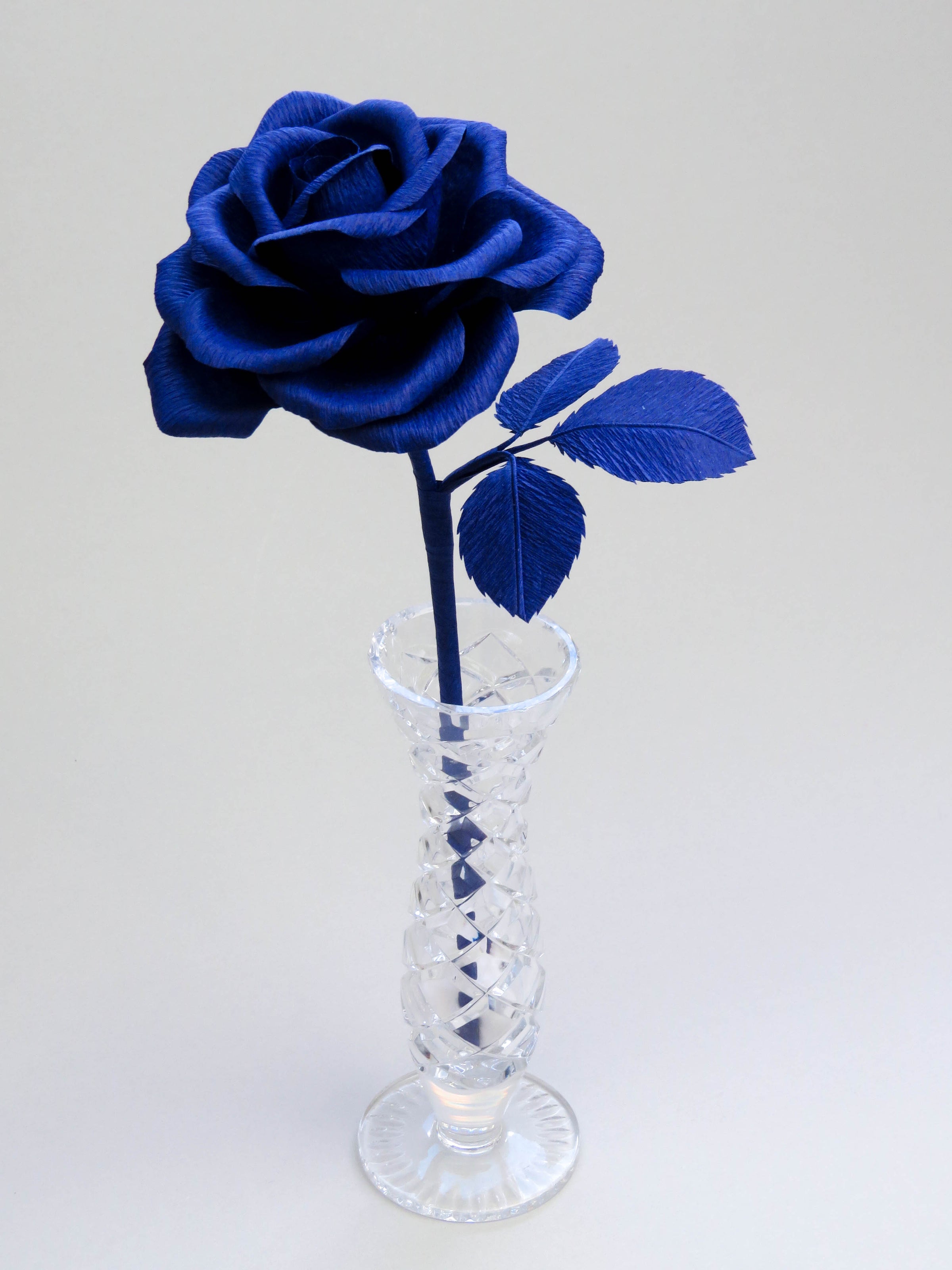 Sapphire blue crepe paper rose with three sapphire blue leaves standing in a narrow glass vase against a light grey backdrop