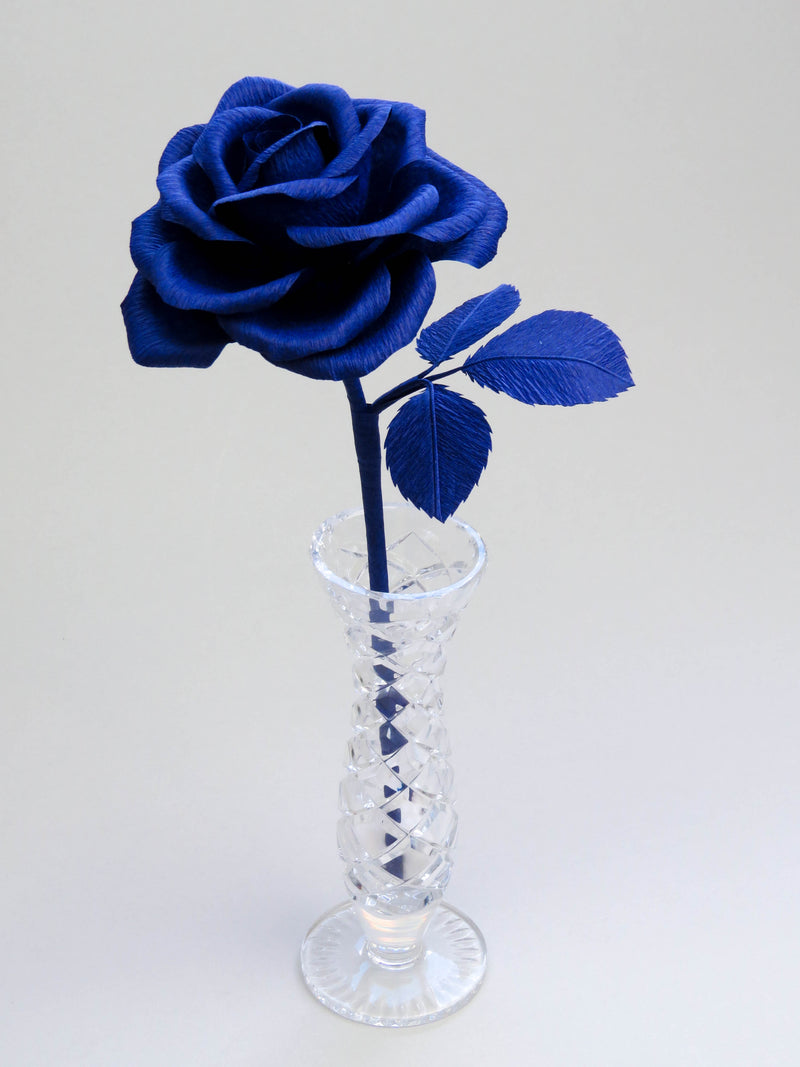 Sapphire blue crepe paper rose with three sapphire blue leaves standing in a narrow glass vase against a light grey backdrop