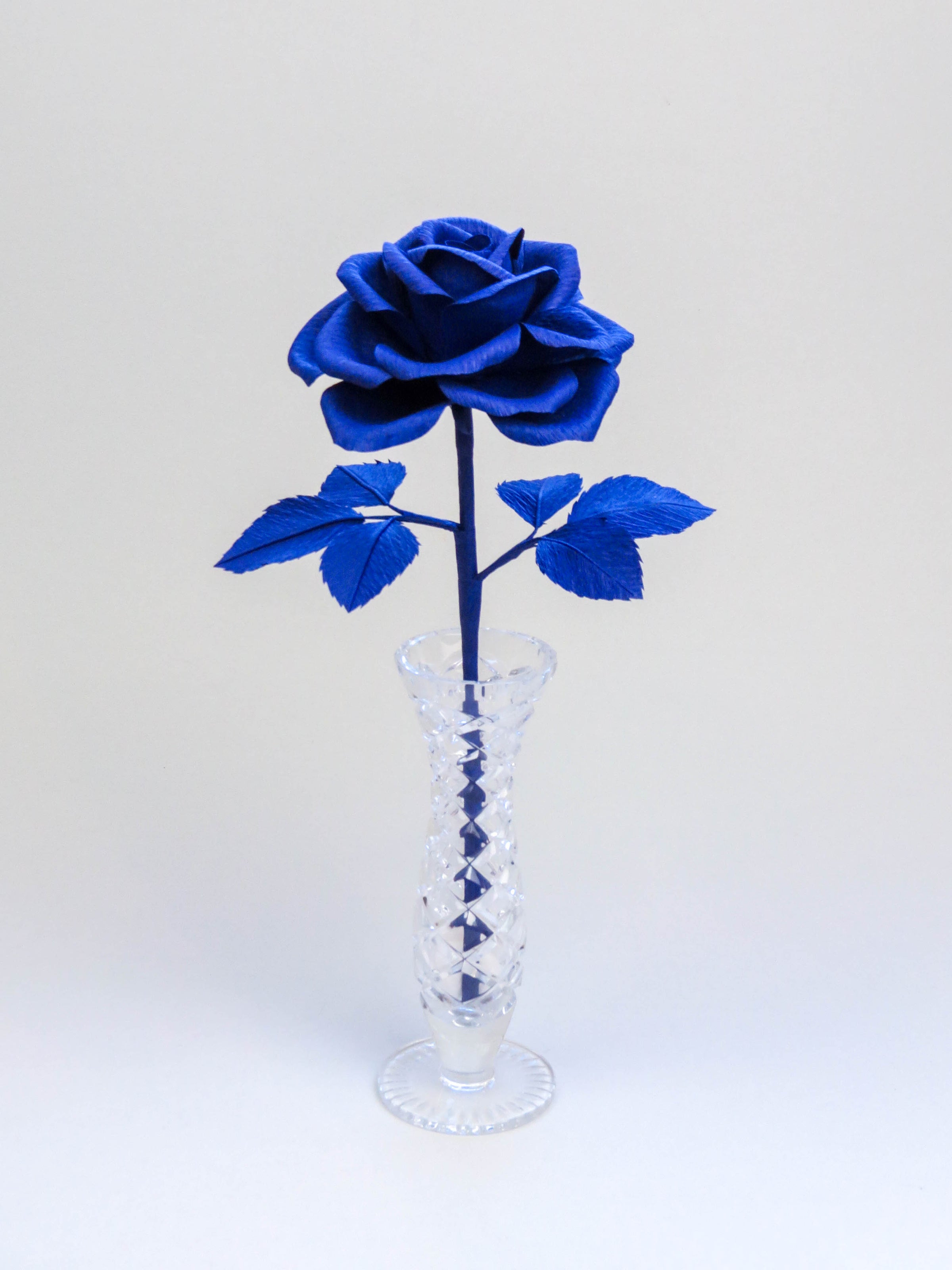 Sapphire blue crepe paper rose with six sapphire blue leaves standing in a narrow glass vase set against a light grey background
