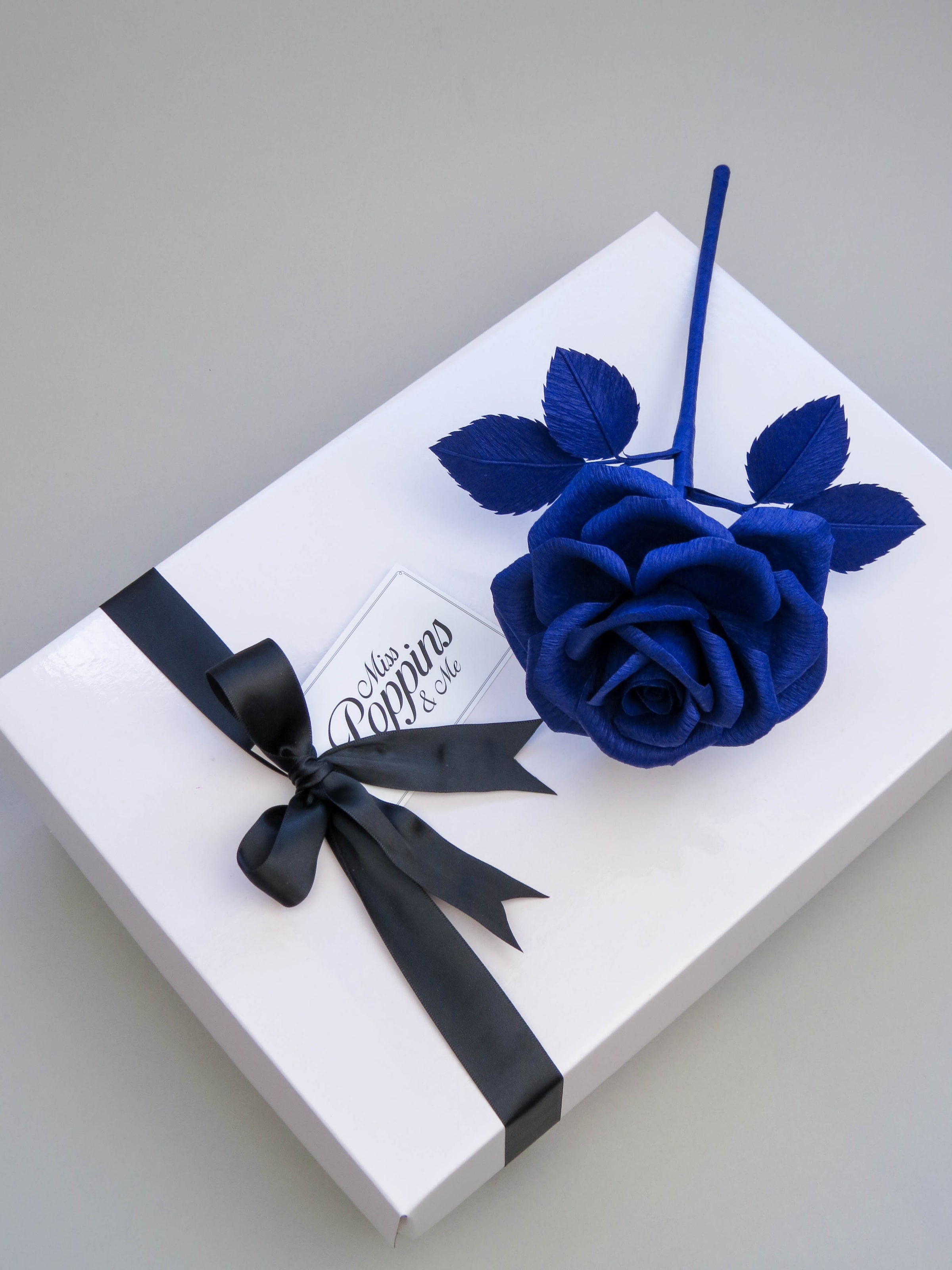 Sapphire blue crepe paper rose lying diagonally on top of a luxury white gift box that has a black satin ribbon tied in a bow with a Miss Poppins and Me gift tag attached