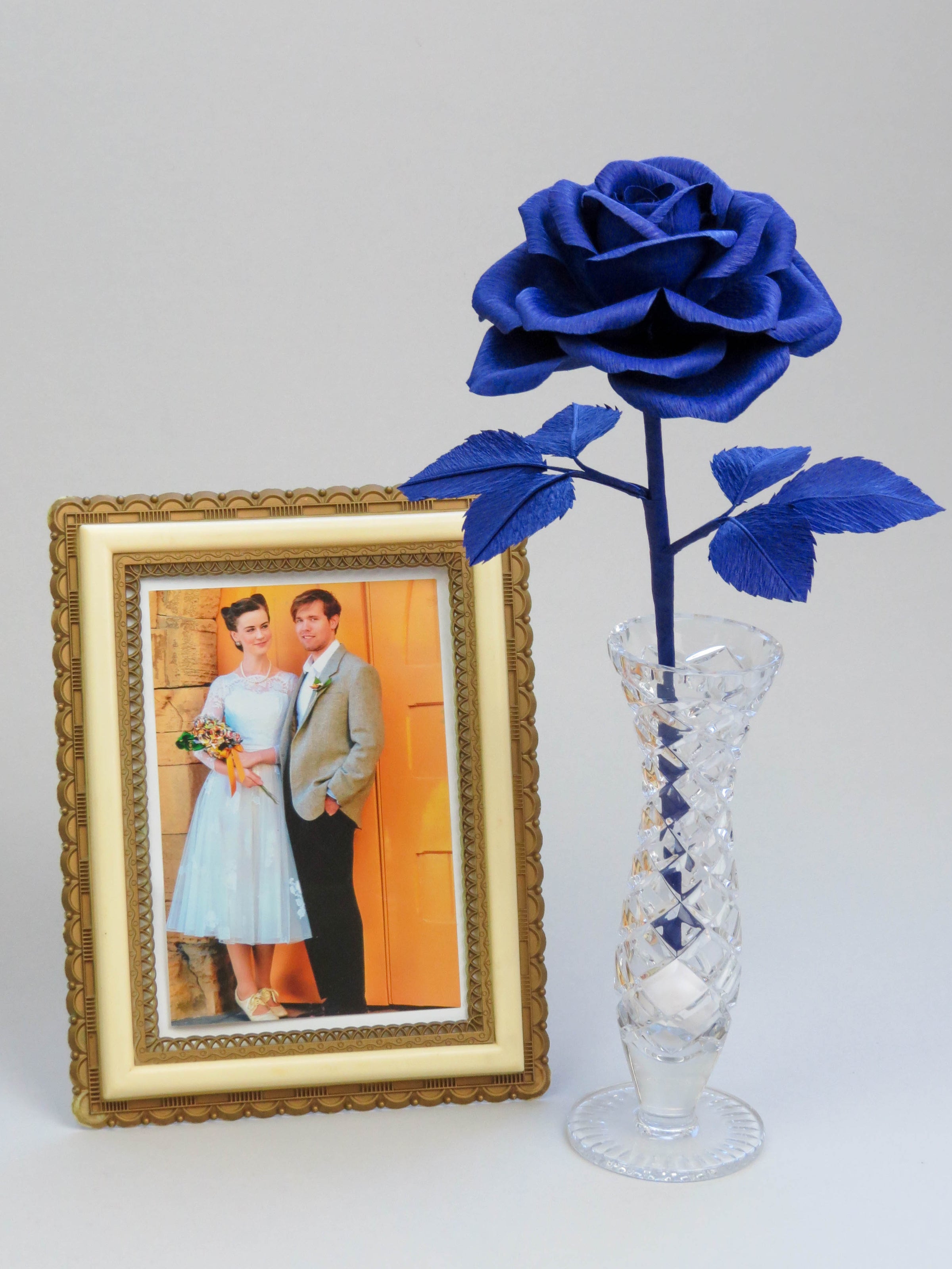 Sapphire blue crepe paper rose with six sapphire blue leaves standing in a slender glass vase with a vintage cream and gold framed wedding photo standing beside it of a bride wearing a 1950s white wedding dress and holding a bouquet of paper flowers, with a groom in a grey jacket and black trousers standing to her right