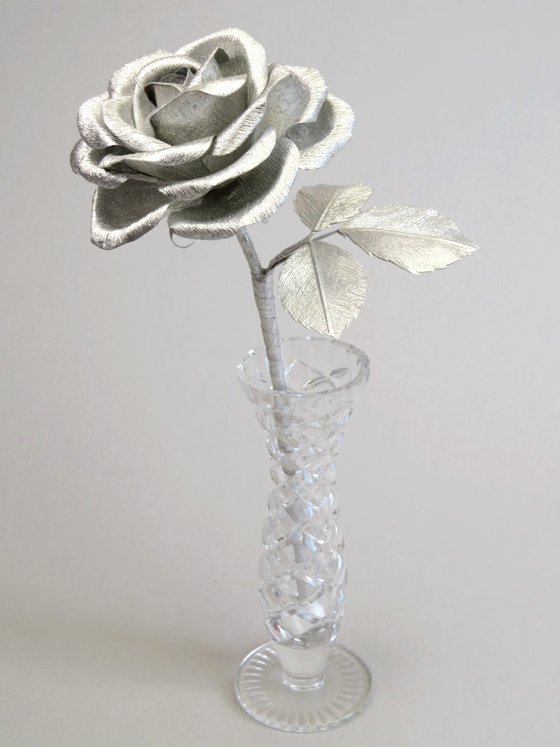 Silver crepe paper rose with three silver leaves standing in a narrow glass vase against a light grey backdrop