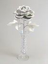 Silver crepe paper rose with six silver leaves standing in a narrow glass vase set against a light grey background