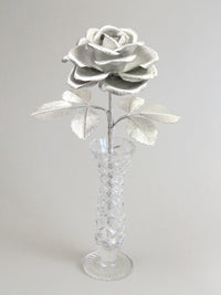 Silver crepe paper rose with six silver leaves standing in a narrow glass vase set against a light grey background