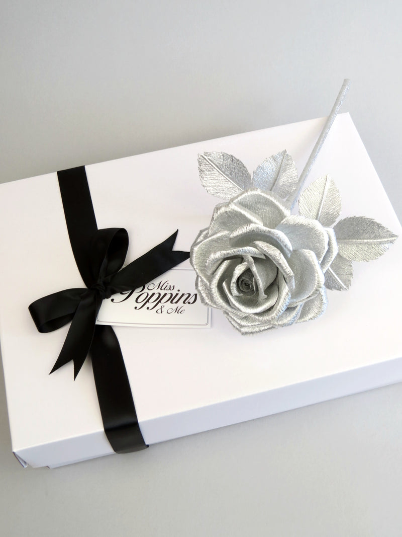Silver crepe paper rose lying diagonally on top of a luxury white gift box that has a black satin ribbon tied in a bow with a Miss Poppins and Me gift tag attached