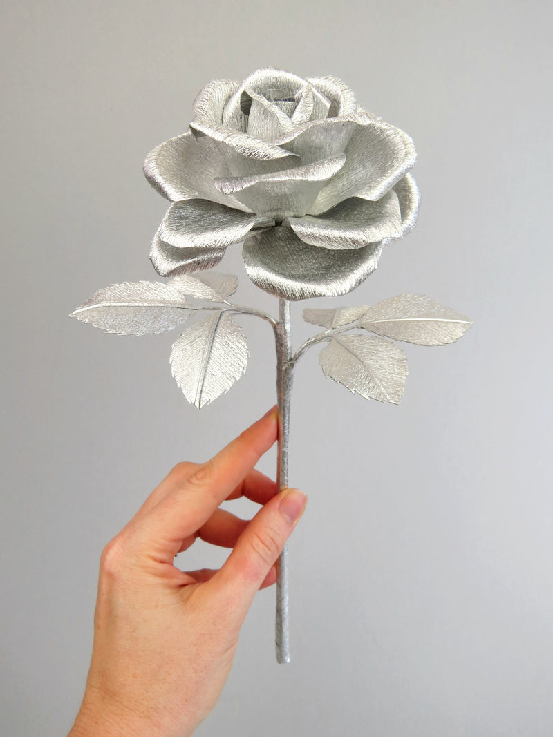 Pale white hand delicately holding the stem of a silver paper rose with six silver leaves