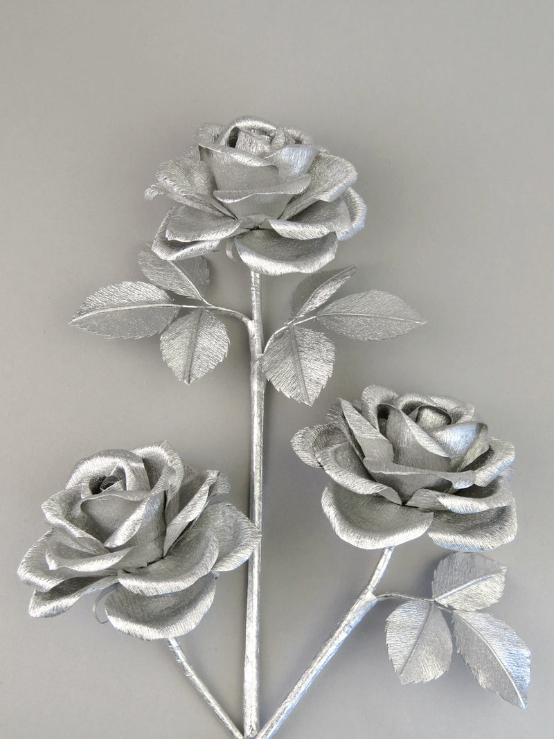 Three silver crepe paper roses randomly lying next to each other on a light grey background. The left rose is leafless, the middle rose has six silver leaves attached and the right rose has three leaves.