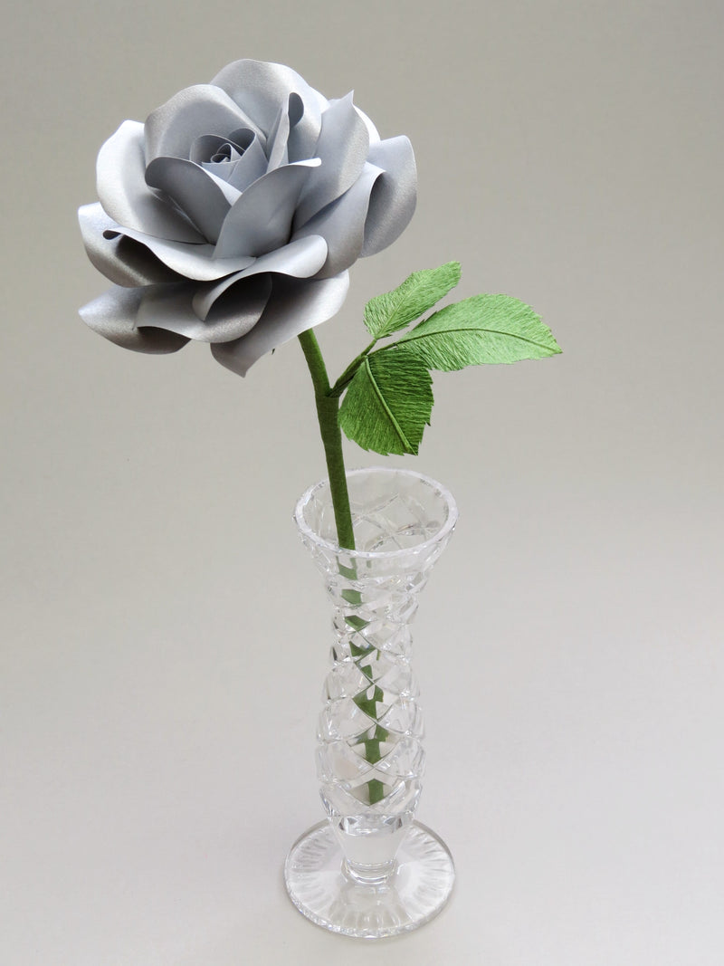 Steel paper rose with three ivy green crepe paper leaves standing in a narrow glass vase against a light grey backdrop