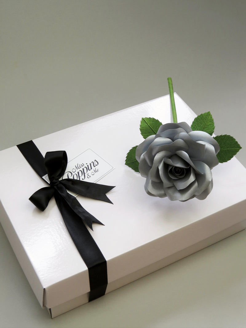 Metallic steel paper rose lying diagonally on top of a luxury white gift box that has a black satin ribbon tied in a bow with a Miss Poppins and Me gift tag attached
