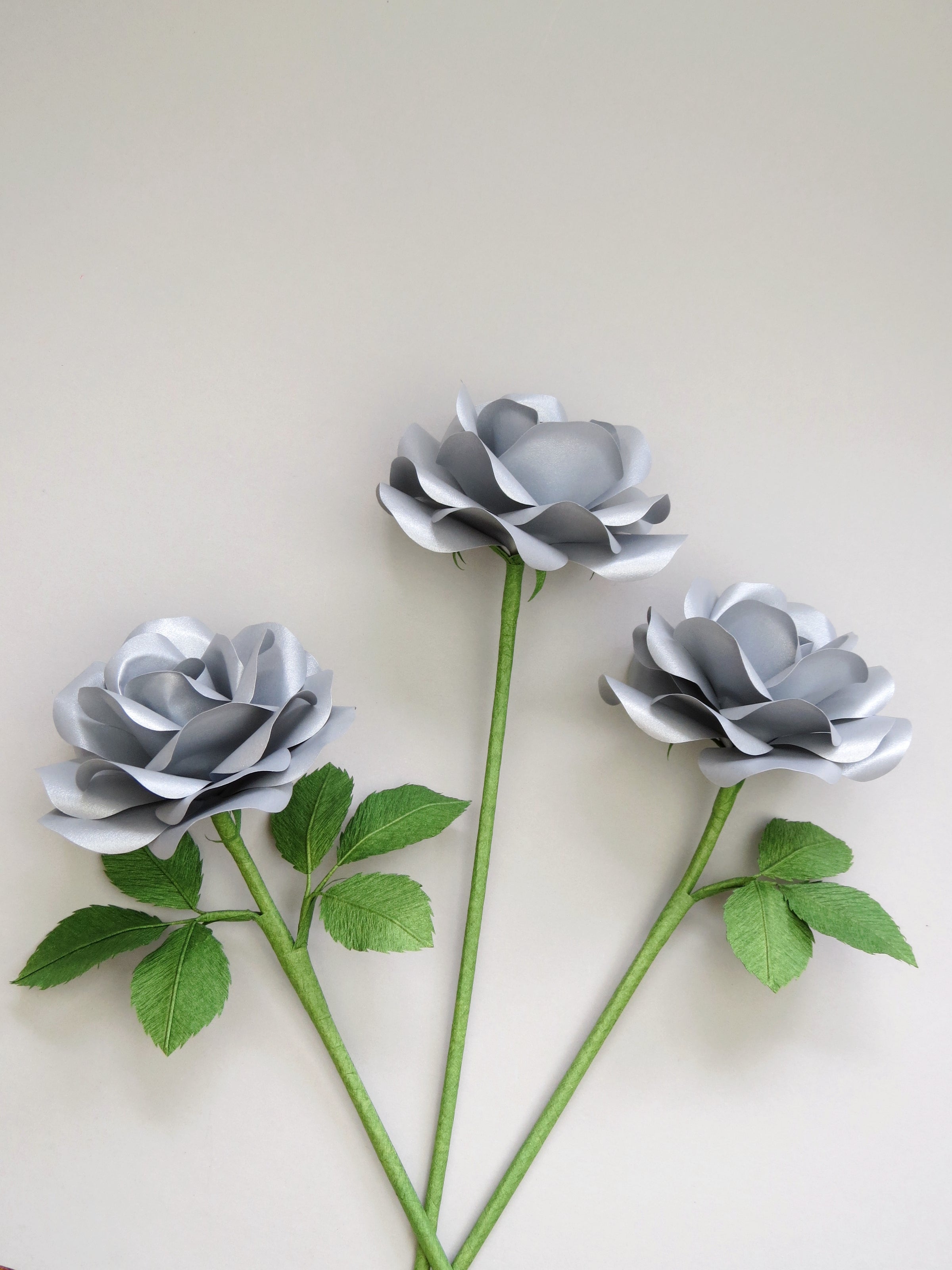 Three metallic steel paper roses randomly lying next to each other on a light grey background. The left rose has six ivy green leaves attached, the middle rose is leafless, the middle and the right rose has three ivy green leaves.