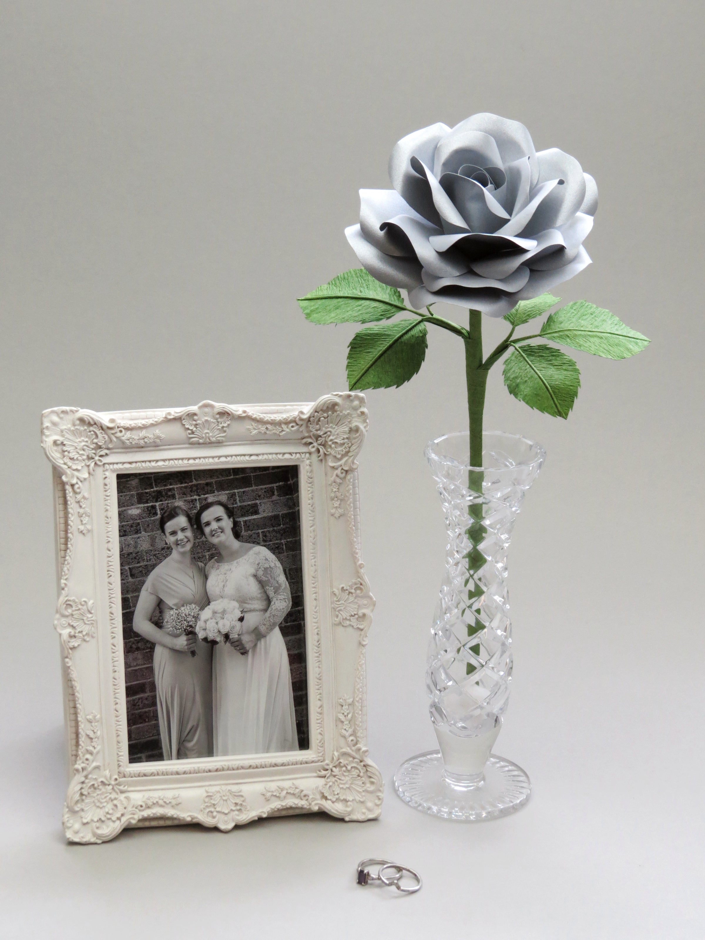 Steel paper rose with six ivy green leaves standing in a slender glass vase with a thick vintage photo frame with a black and white photo of a happy bride and her bridesmaid, and a platinum wedding and engagement ring delicately placed on top of each other nearby