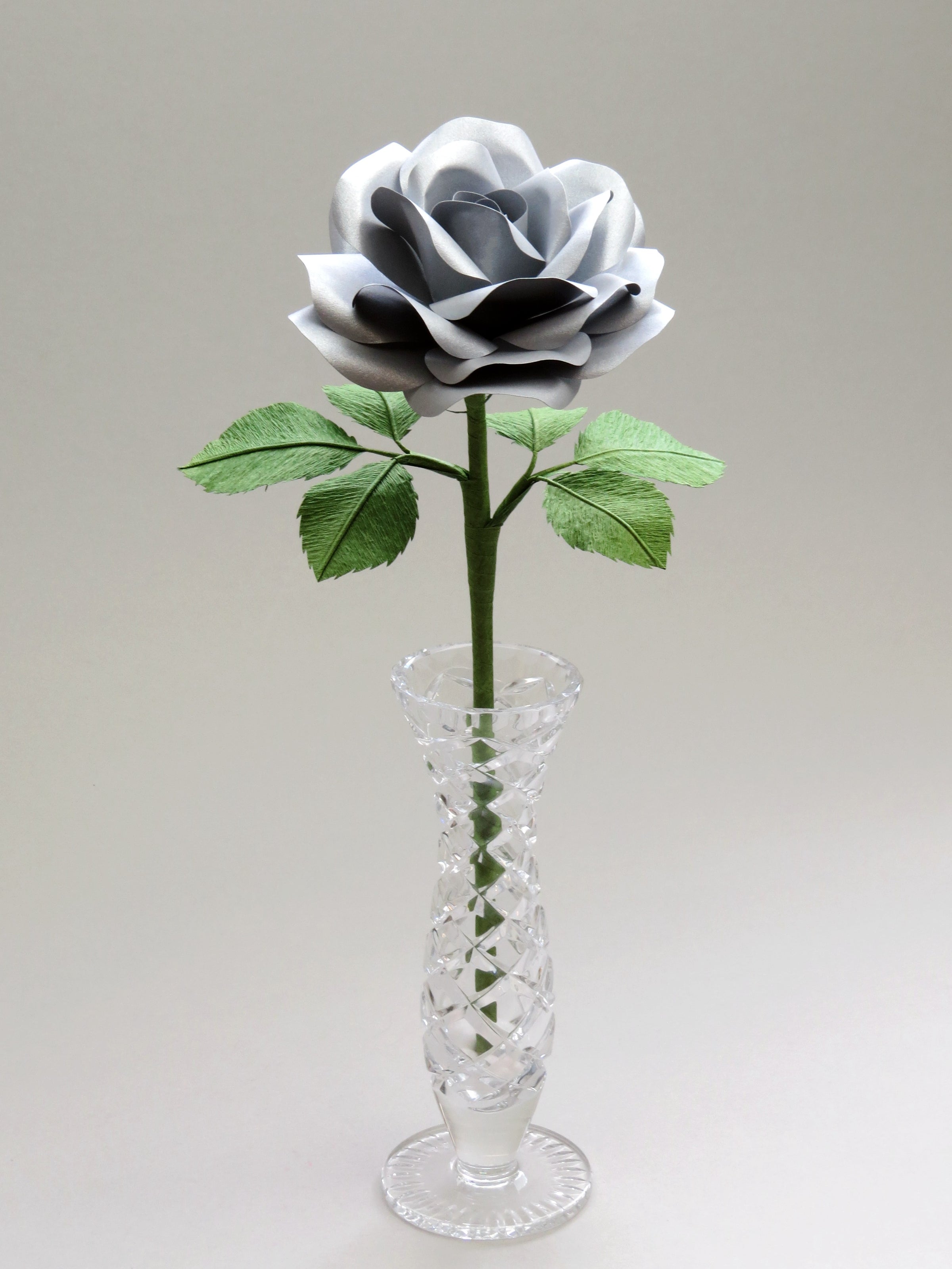 Steel paper rose with six ivy green crepe paper leaves standing in a narrow glass vase set against a light grey background