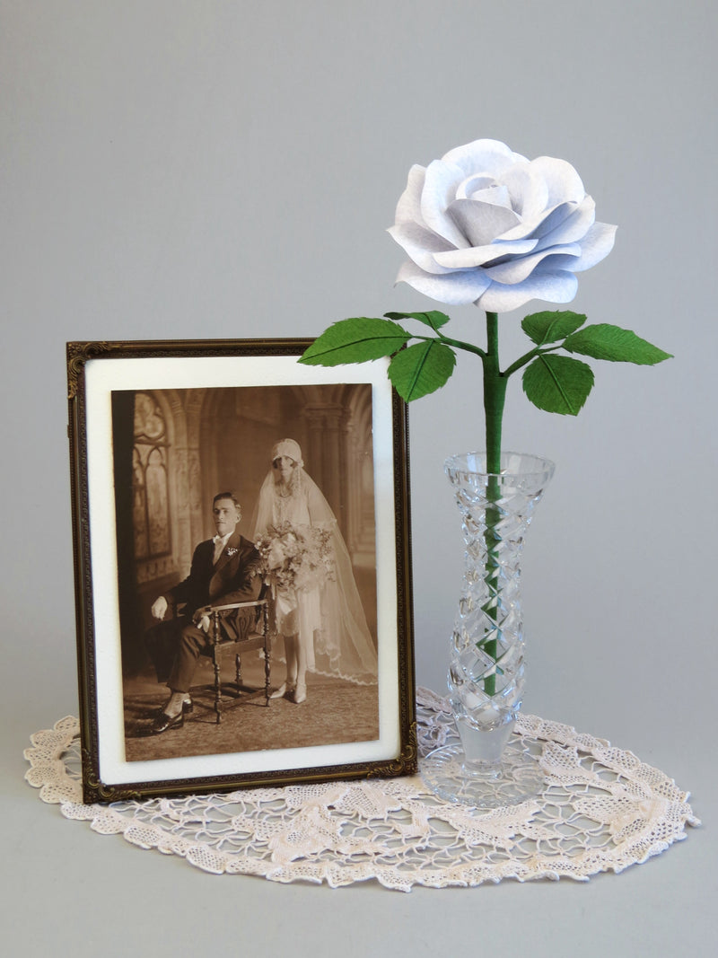 White lace printed paper rose with six ivy green leaves standing in a slender glass vase with a thin gold framed sepia wedding photo of a 1920’s bride and groom standing beside it, all sitting on top of an ivory oval doily