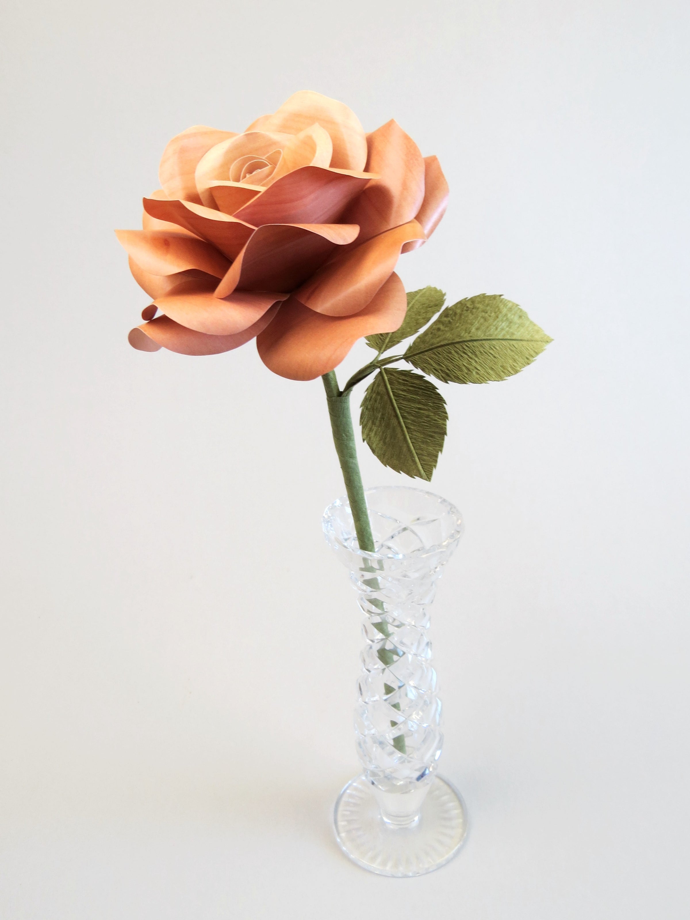Light brown willow printed paper rose with three olive green leaves standing in a narrow glass vase against a light grey backdrop