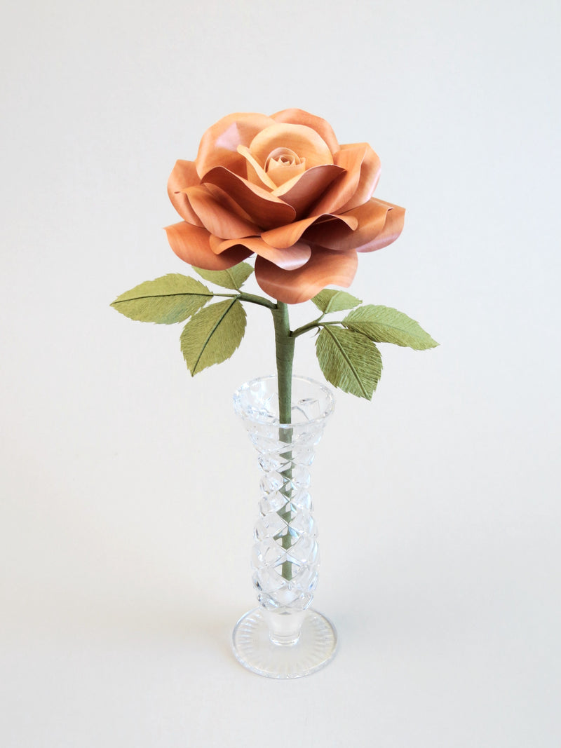 Light brown willow printed paper rose with six olive green leaves standing in a narrow glass vase set against a light grey background