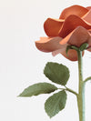 The underneath of the light brown willow printed paper rose showing the olive green calyx, with olive green stem and the back of the 3 olive green crepe paper leaves