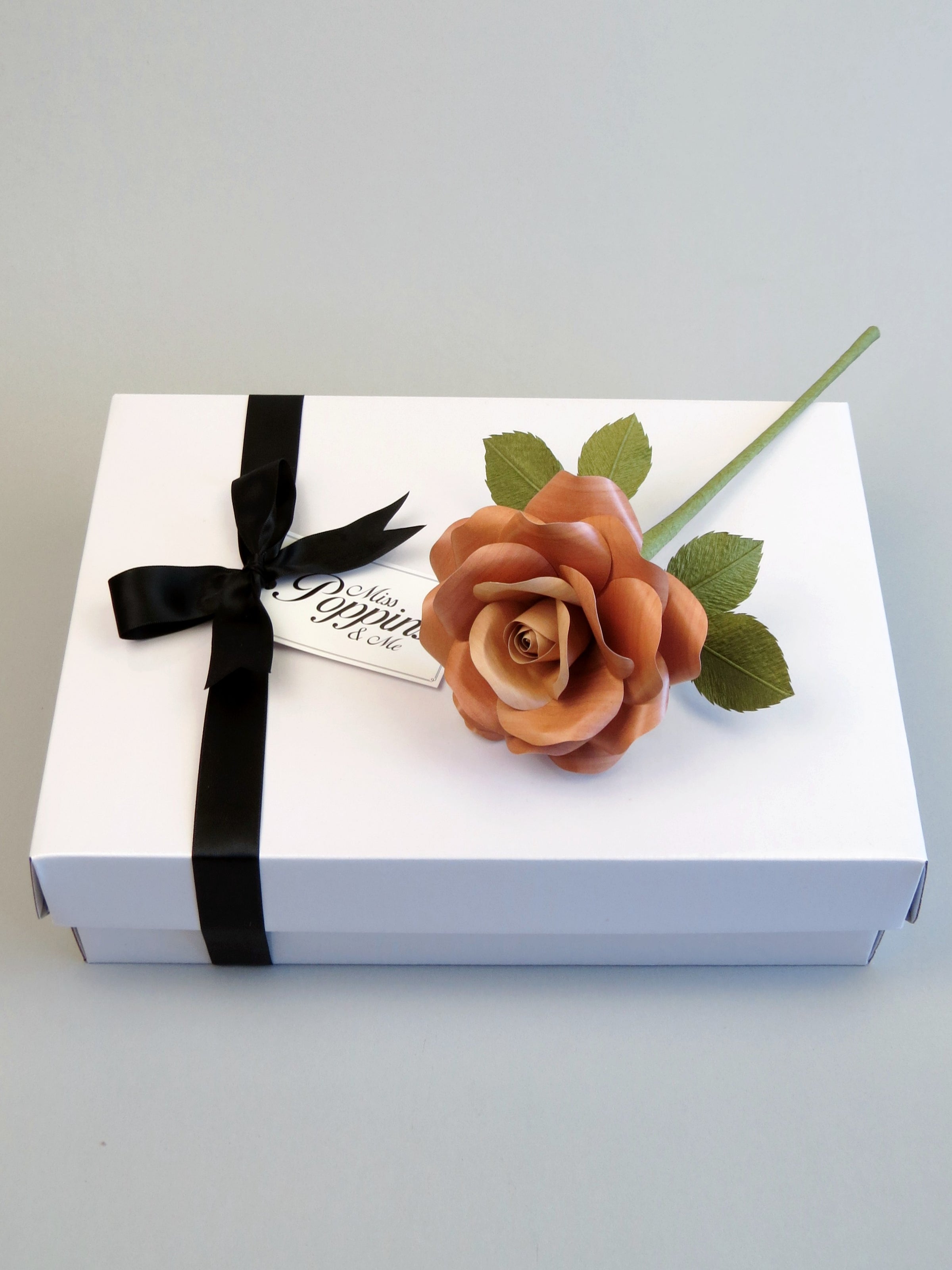 Light brown willow printed paper rose lying on top of a luxury white gift box that has a black satin ribbon tied in a bow with a Miss Poppins and Me gift tag attached