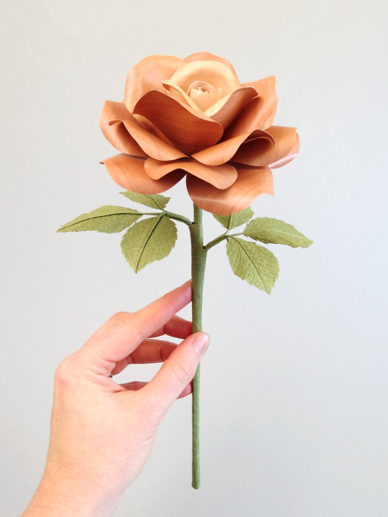 Pale white hand delicately holding the stem of a light brown willow printed paper rose with six olive green leaves