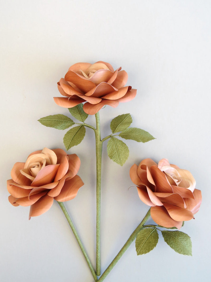 Three light brown willow printed paper roses randomly lying next to each other on a light grey background. The left rose has no leaves, the middle rose has six olive green leaves and the right rose has three olive green leaves.