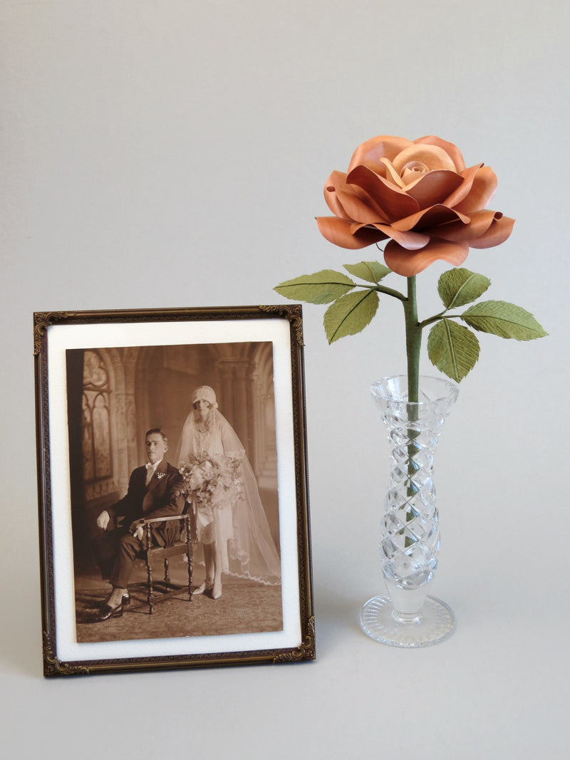 Light brown willow printed paper rose with six olive green leaves standing in a slender glass vase to the right of a vintage framed sepia wedding photo of a 1920’s seated groom with bride standing behind him