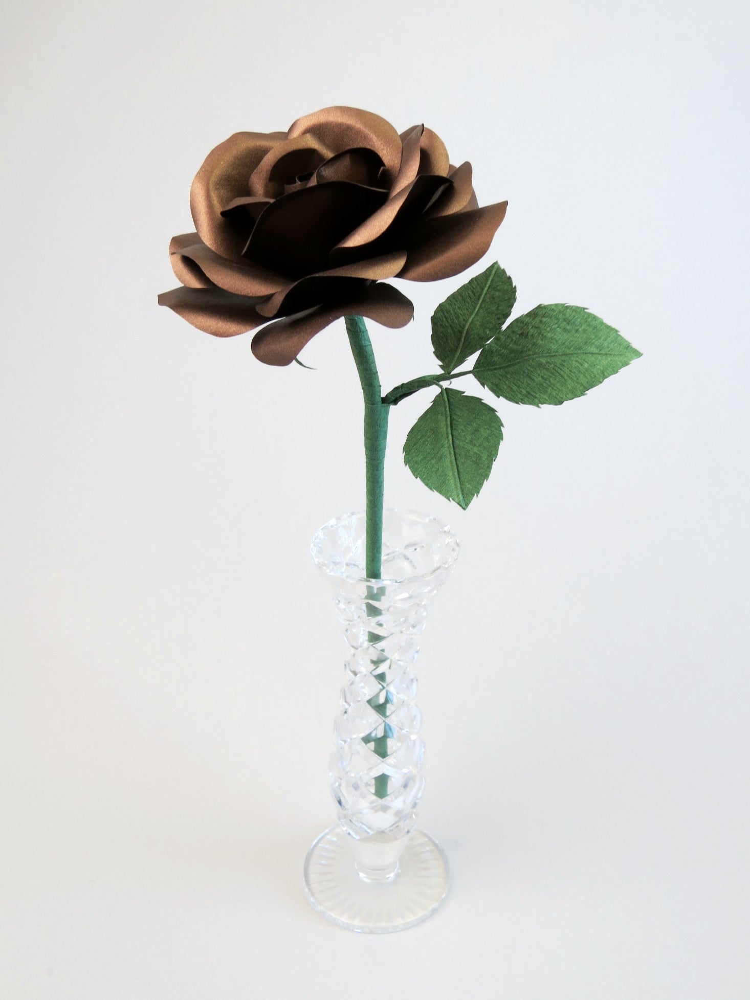 Bronze paper rose with three leaves standing in a narrow glass vase against a light grey backdrop