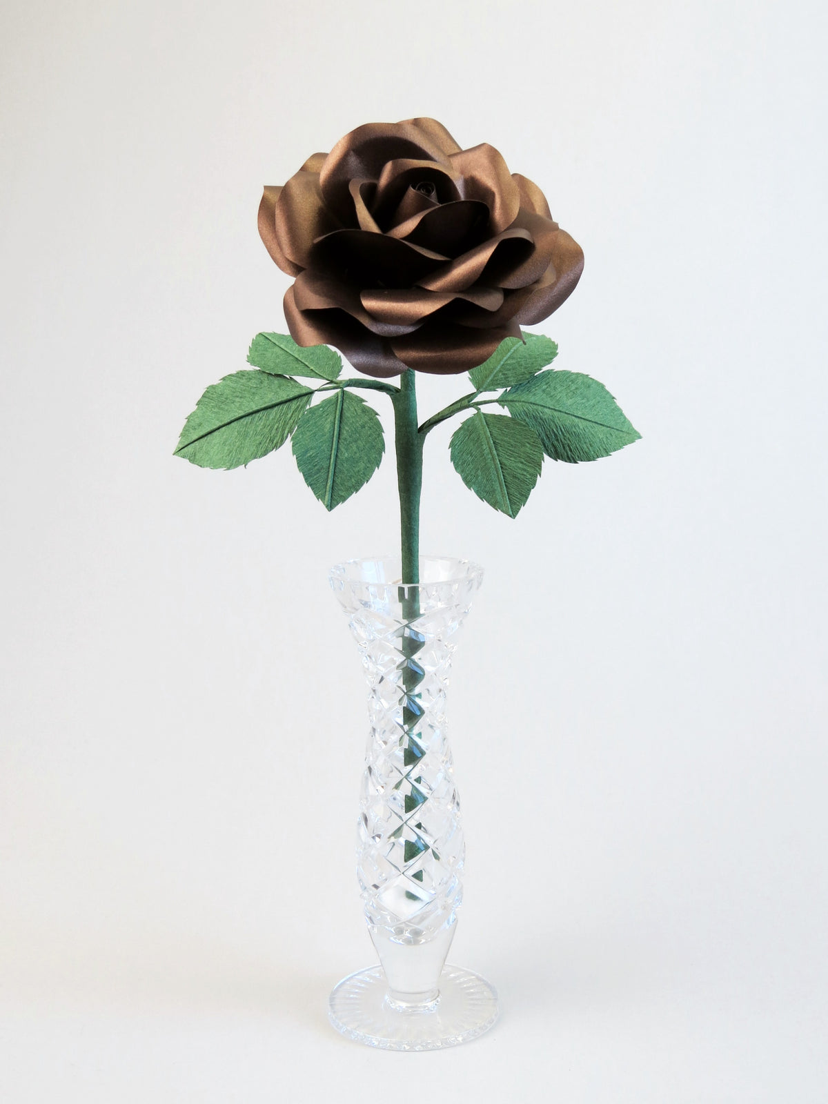 Bronze paper rose with six forest green leaves standing in a narrow glass vase set against a light grey background