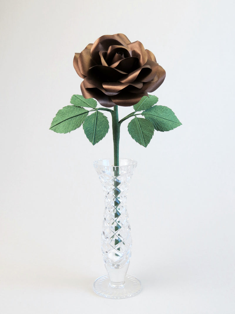 Bronze paper rose with six forest green leaves standing in a narrow glass vase set against a light grey background