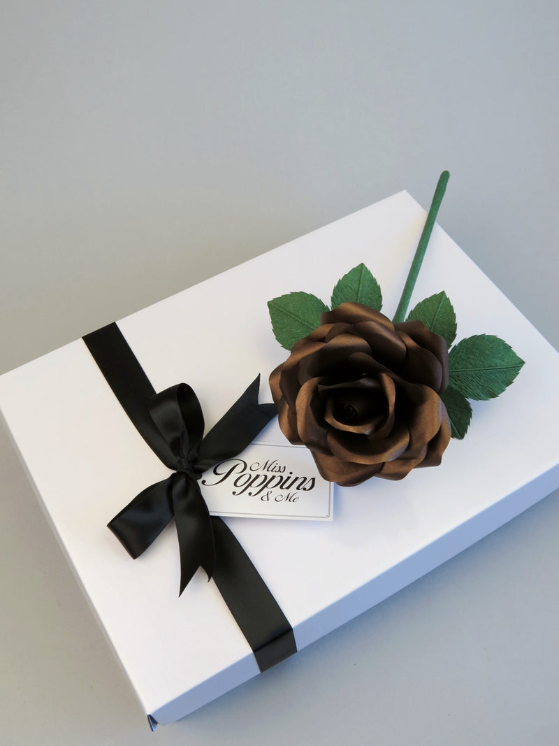 Bronze paper rose lying on top of a luxury white gift box that has a black satin ribbon tied in a bow with a Miss Poppins and Me gift tag attached