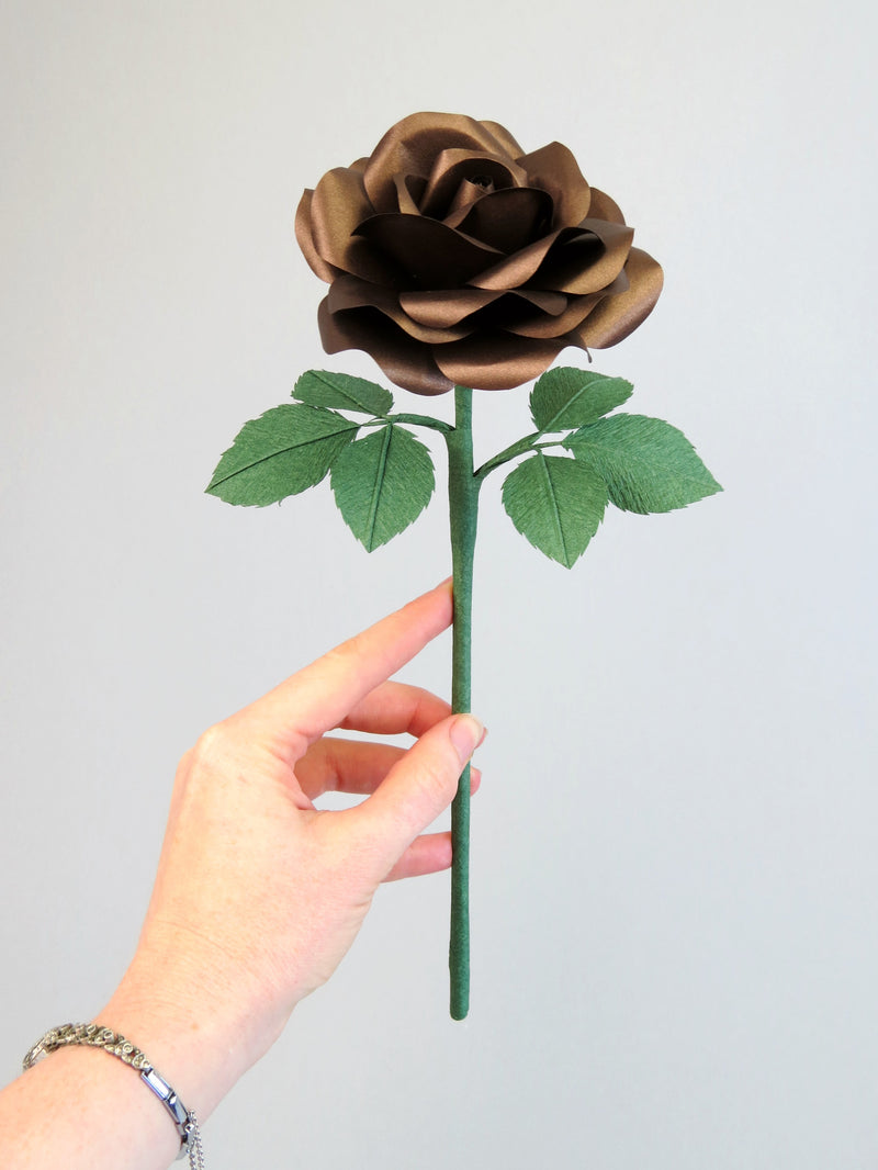 Pale white hand delicately holding the stem of a bronze paper rose with six leaves