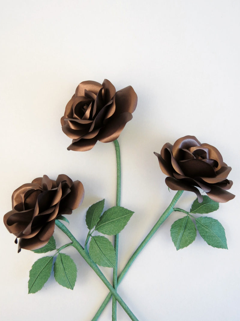 Three bronze paper roses randomly lying next to each other on a light grey background. The left rose has six forest green leaves attached, the middle rose has no leaves and the right rose has three leaves.