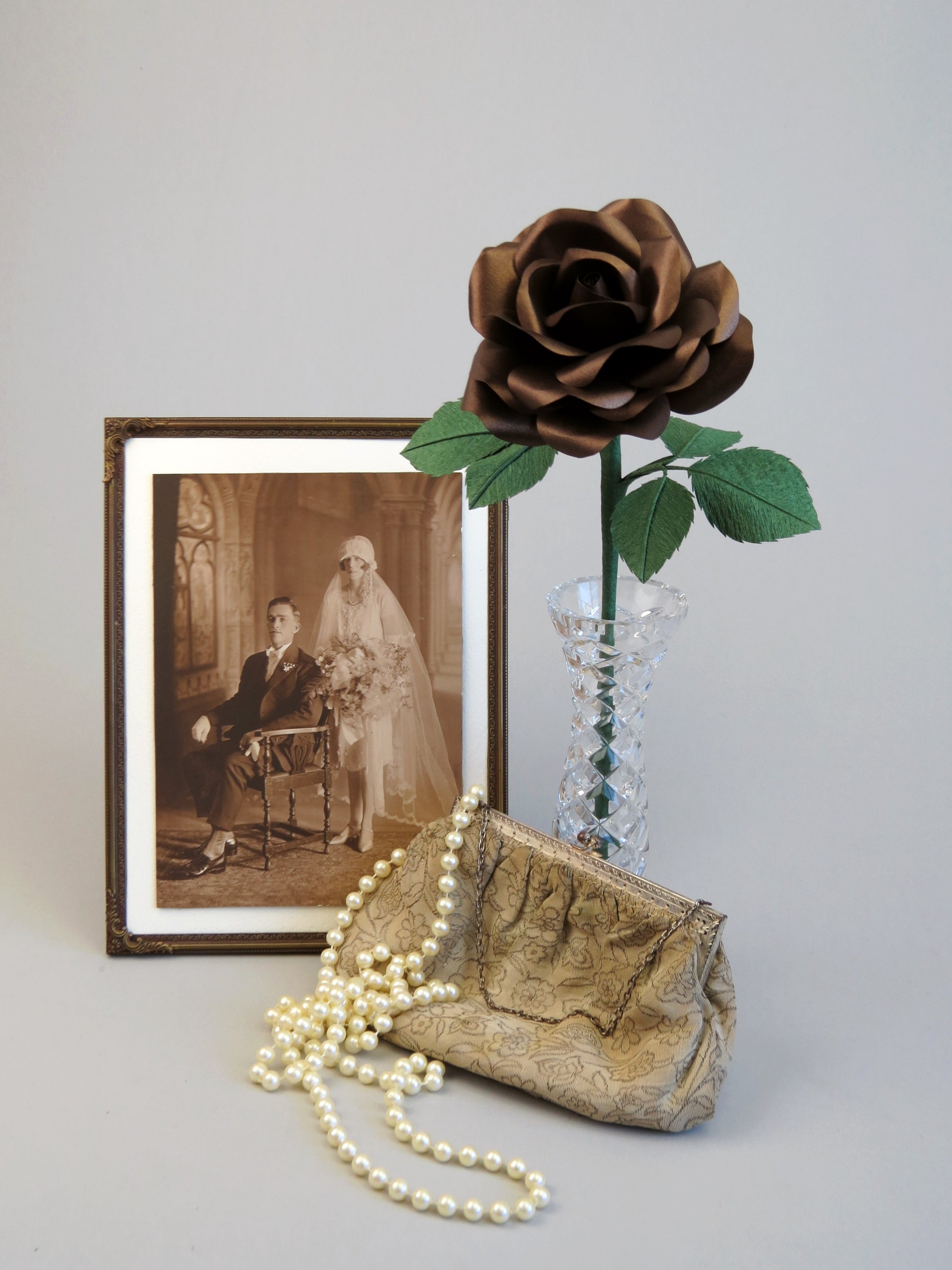 Bronze paper rose with six green leaves standing in a slender glass vase with a framed vintage wedding photo of a 1920’s bride and groom standing to the left of the vase. A cream and metallic vintage clutch rests against the front base of the vase with a long pearl necklace draped over it. 