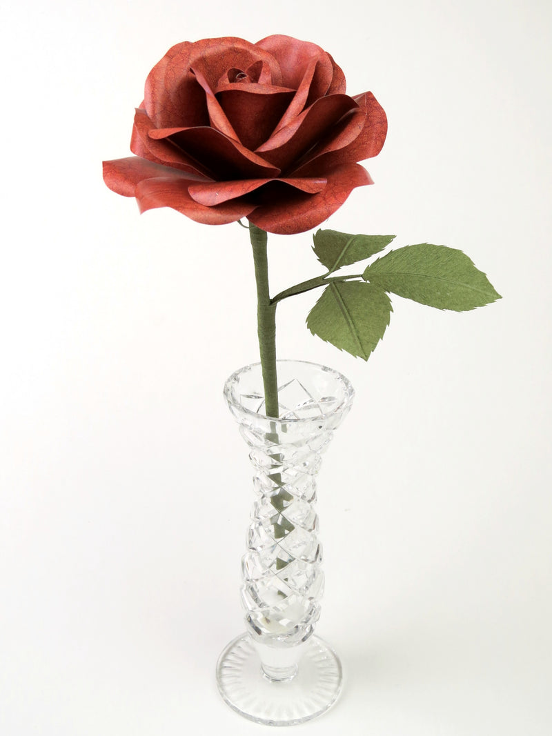Brown leather grain paper rose with three leaves standing in a narrow glass vase against a light grey backdrop