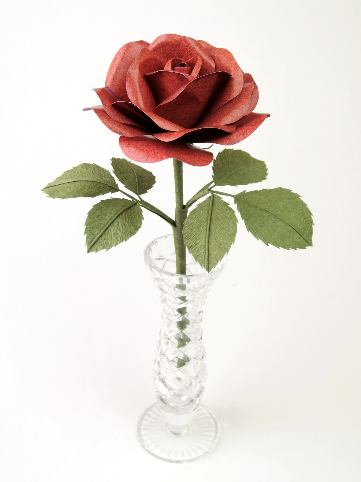 Brown leather grain paper rose with six leaves standing in a narrow glass vase against a light grey background