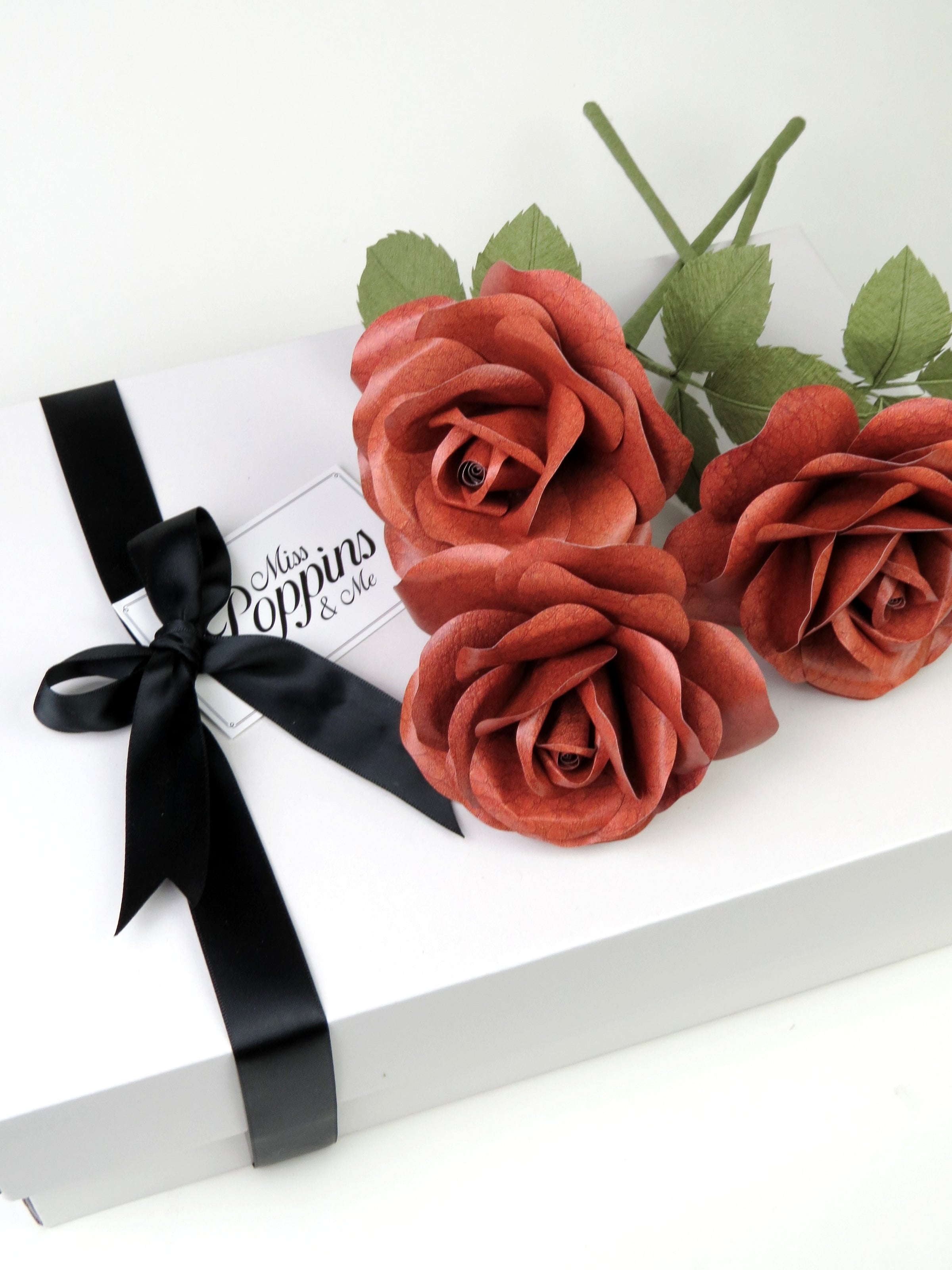 A bouquet of three brown leather grain paper rose lying on top of a luxury white gift box that has a black satin ribbon tied in a bow with a Miss Poppins and Me gift tag attached