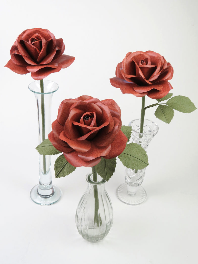 Three brown leather grain paper roses in separate glass vases. The left rose has no leaves, the middle rose has six leaves and the third rose has three leaves. 