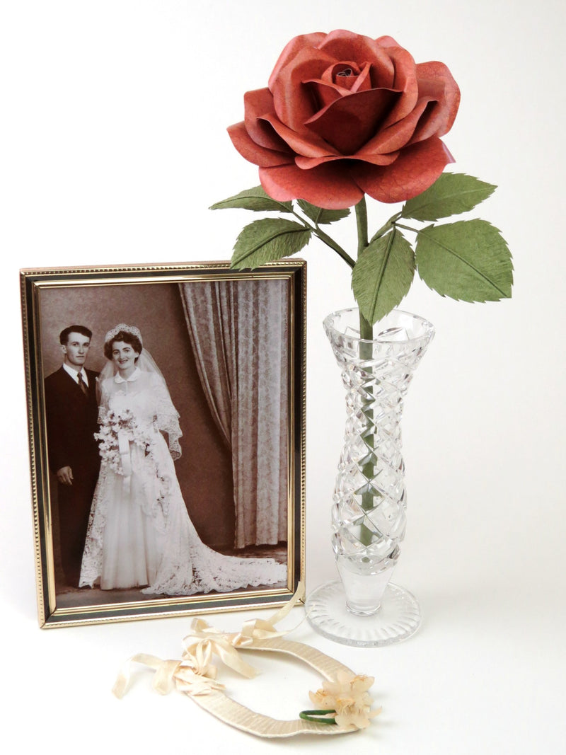 Brown leather grain paper rose with six green leaves standing in a slender glass vase with an old framed sepia wedding photo standing beside it and a vintage cream ribboned horseshoe lying in front 