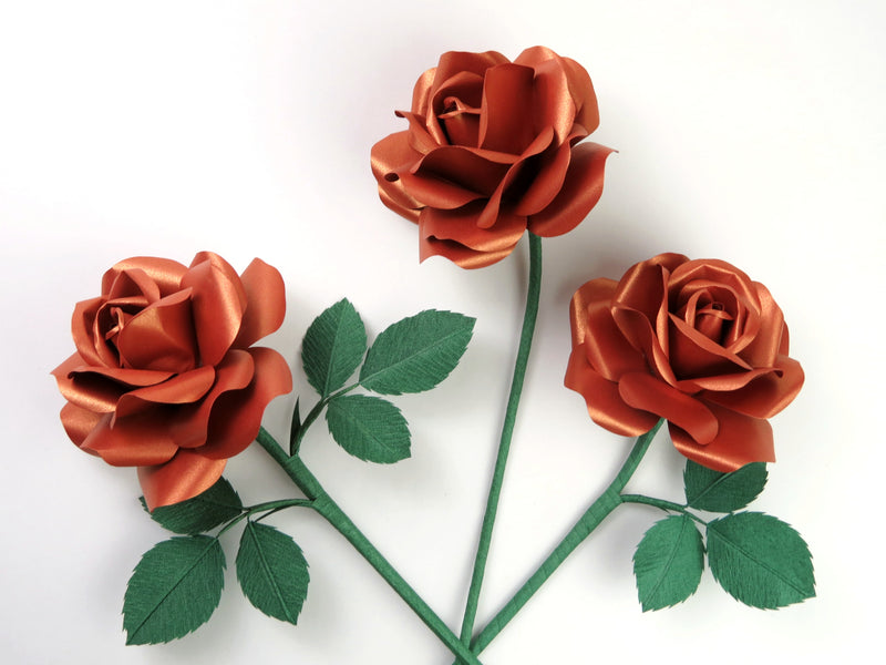 Three copper paper roses randomly lying next to each other on a light grey background. The left rose has six forest green leaves attached, the middle rose has no leaves and the right rose has three leaves.