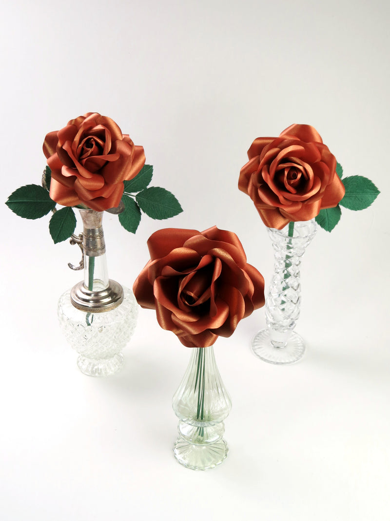 Three copper paper roses each standing in a mismatched glass vase against a light grey background. The left rose has six green leaves attached, the middle rose has no leaves and the right rose has three leaves.