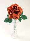 Copper paper rose with six green leaves standing in a narrow glass vase set against a light grey background