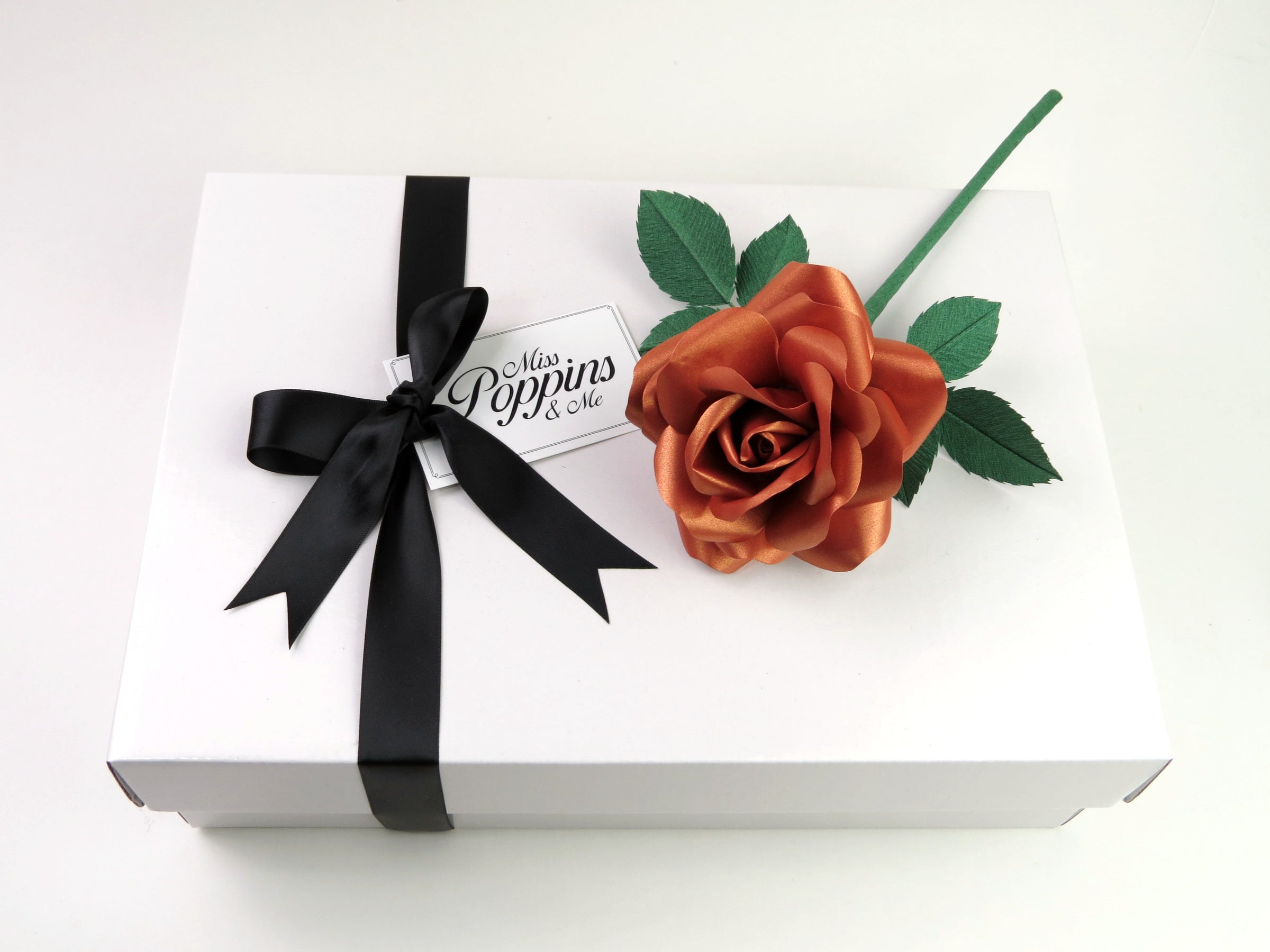 Copper paper rose lying on top of a luxury white gift box that has a black satin ribbon tied in a bow with a Miss Poppins and Me gift tag attached