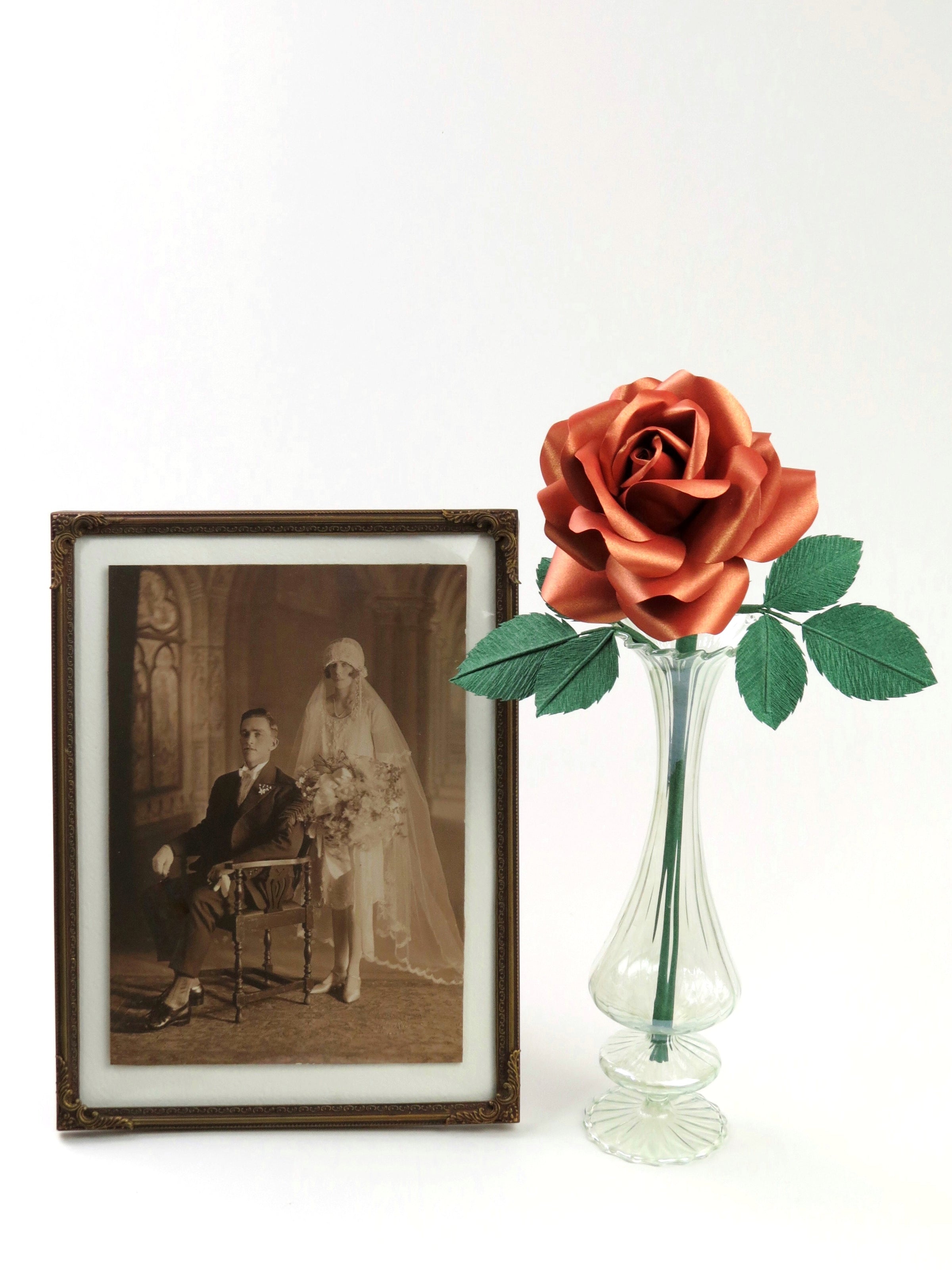 Copper paper rose with six green leaves standing in a slender glass vase with a framed vintage wedding photo of a 1920’s bride and groom standing to the left of the vase
