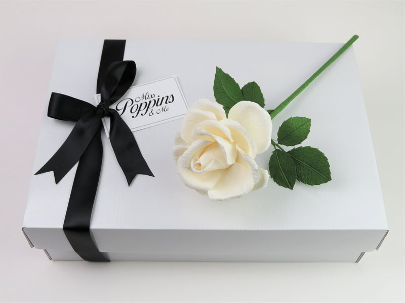 White paper rose lying on top of a luxury white gift box that has a black satin ribbon tied in a bow with a Miss Poppins and Me gift tag attached