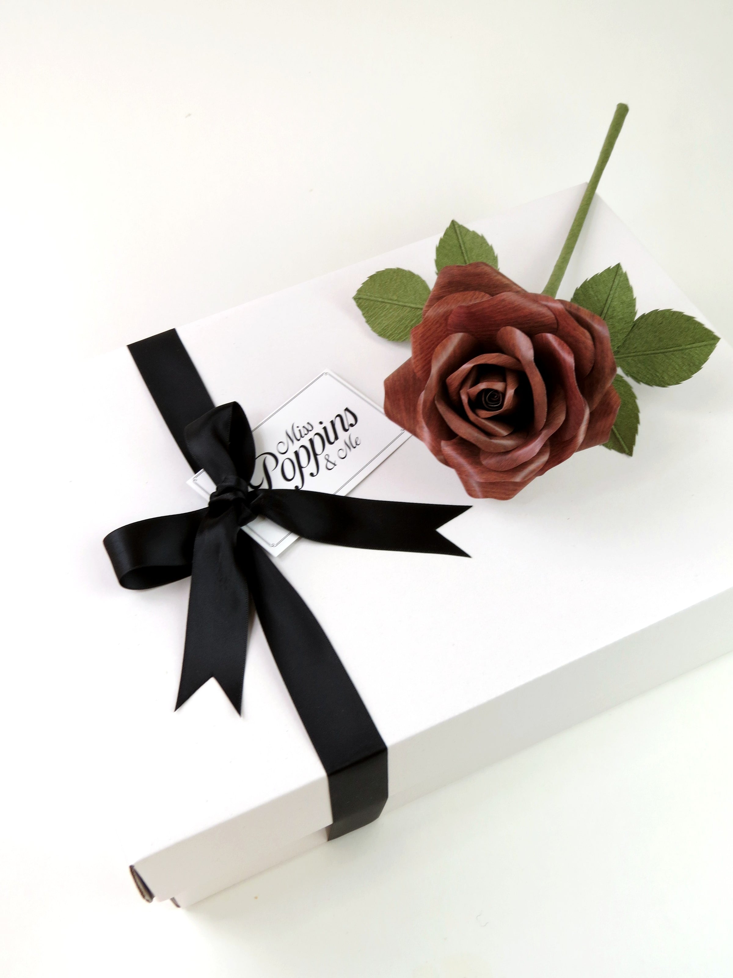 Dark wood grain paper rose lying on top of a luxury white gift box that has a black satin ribbon tied in a bow with a Miss Poppins and Me gift tag attached