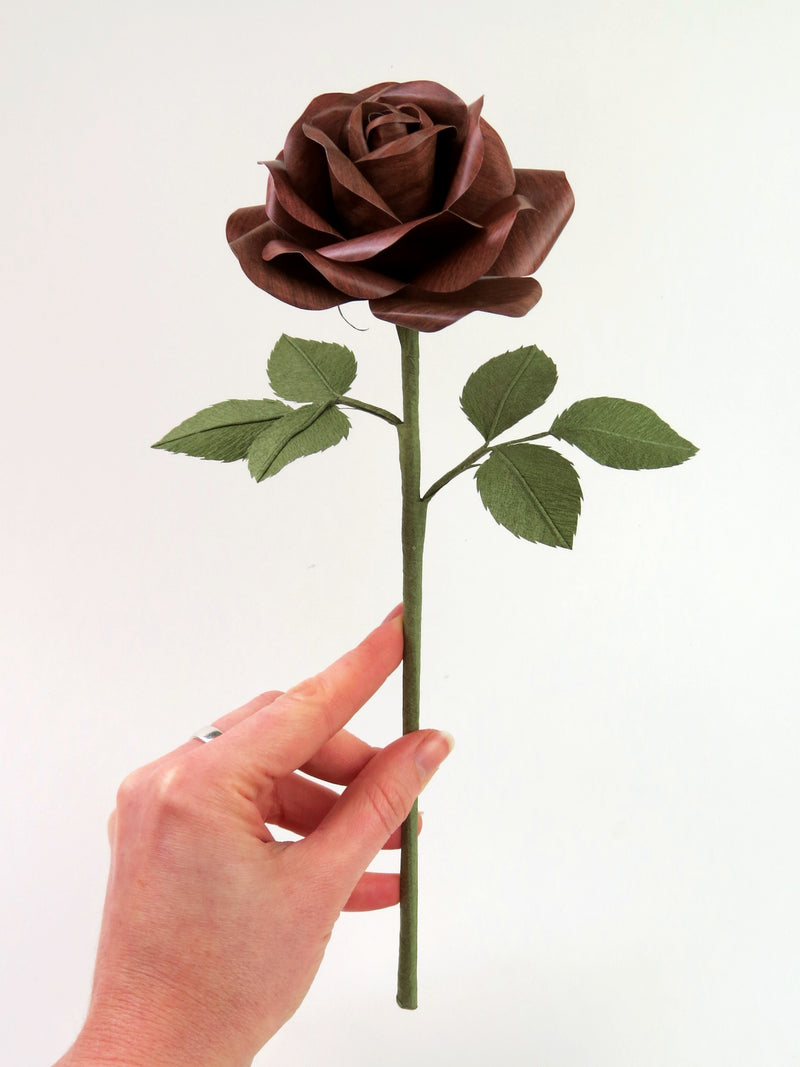 Pale white hand delicately holding the stem of a dark wood grain paper rose with six leaves