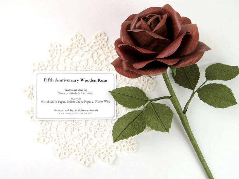 Dark wood grain paper rose lying on a small white vintage doily which has a product tag resting on it detailing the meaning and materials of this paper anniversary rose