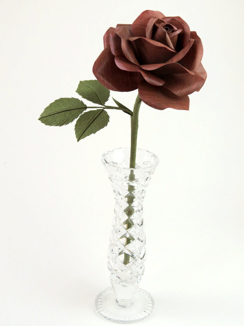 Dark wood grain paper rose with three leaves standing in a narrow glass vase against a white backdrop